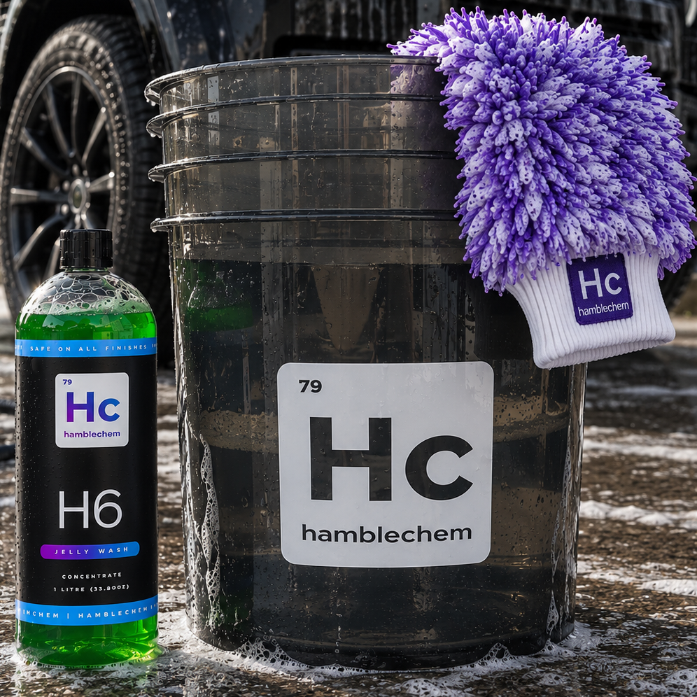 A bucket of Hamblechem H6 car wash concentrate shampoo wash, shown with a bottle of the green liquid concentrate and a purple-and-white microfiber wash mitt branded Hamblechem, set outdoors on wet pavement.